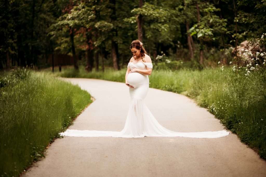 Pregnant woman wearing a white gown posed on a walkway looking down at her baby bump