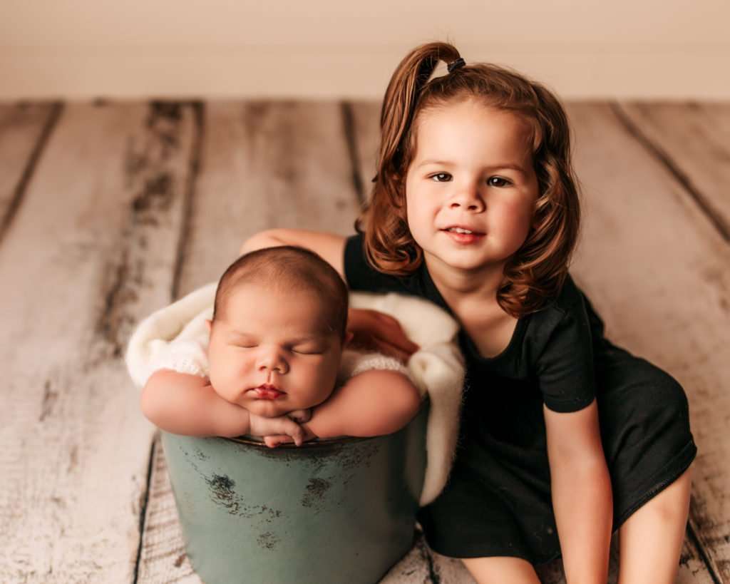 big sister and little brother sitting on wood floor with baby in bucket sleeping and sister smiling at camera for newborn photography images