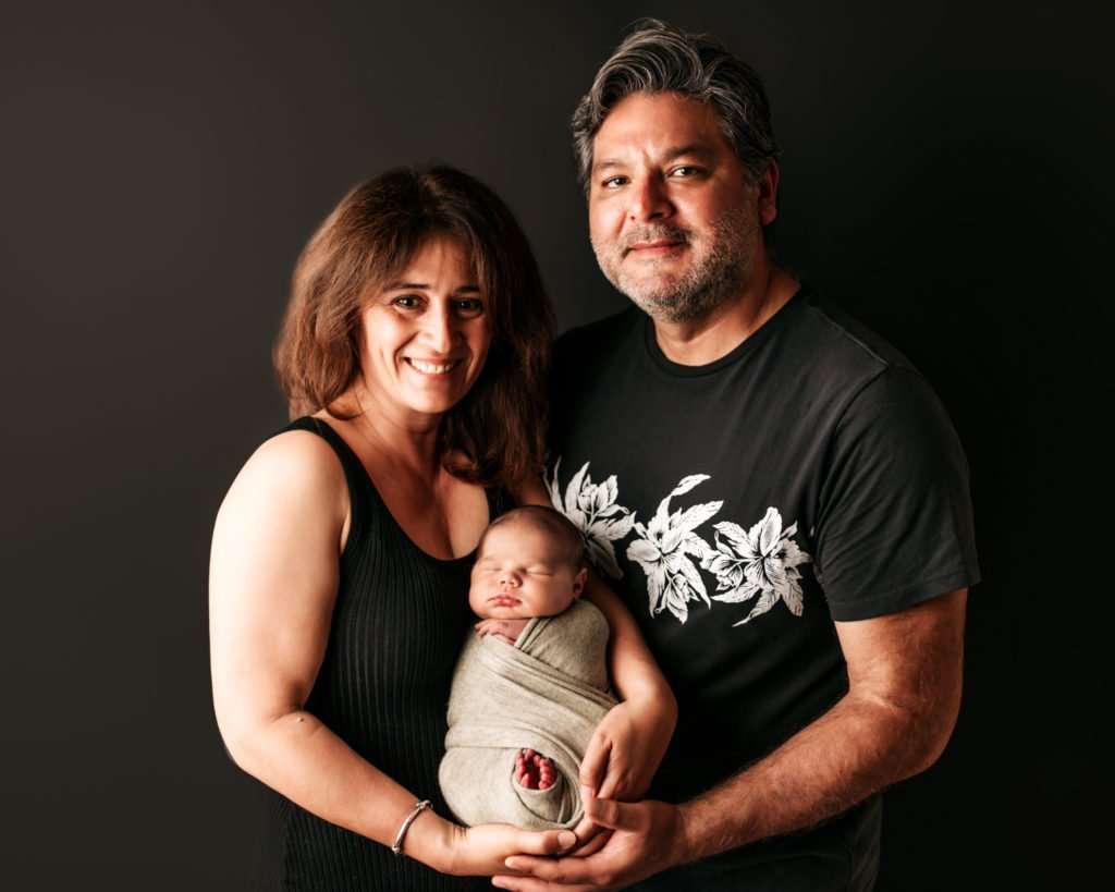 grandparents holding baby against a black backdrop with baby wrapped in their arms for newborn photography images