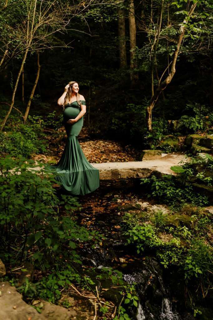 pregnant woman standing on stone bridge in wooded area in dark green dress