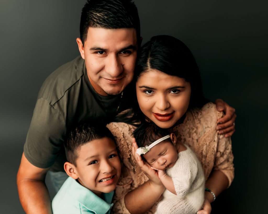 mom holding newborn baby girl with husband and baby's little brother huddled next to them smiling during a newborn photography session