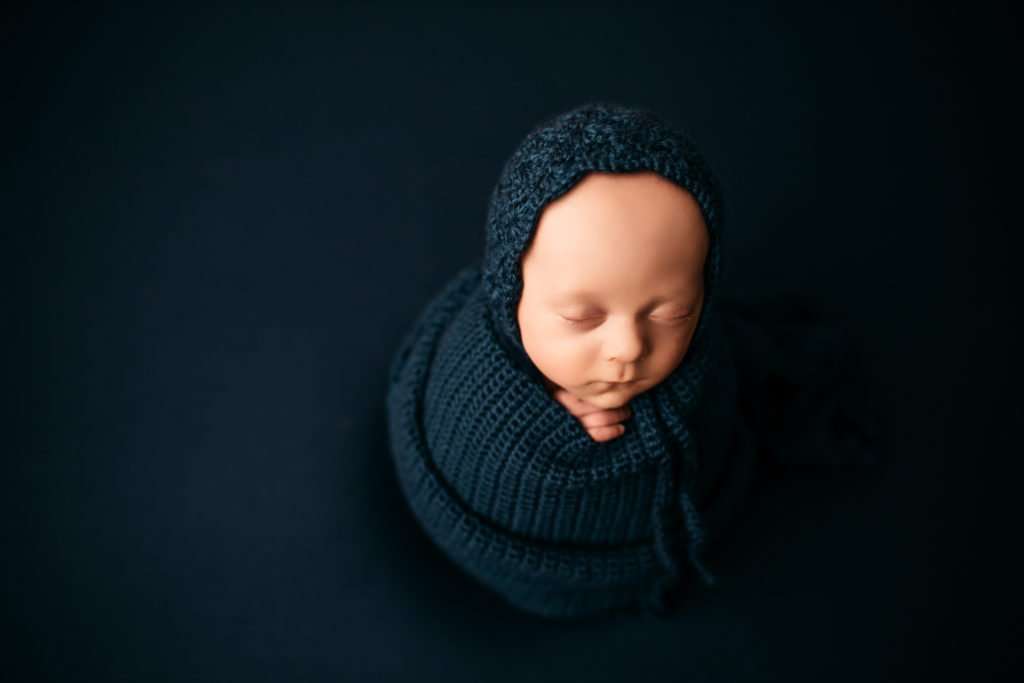 baby wrapped in ball on a blue backdrop with bonnet on