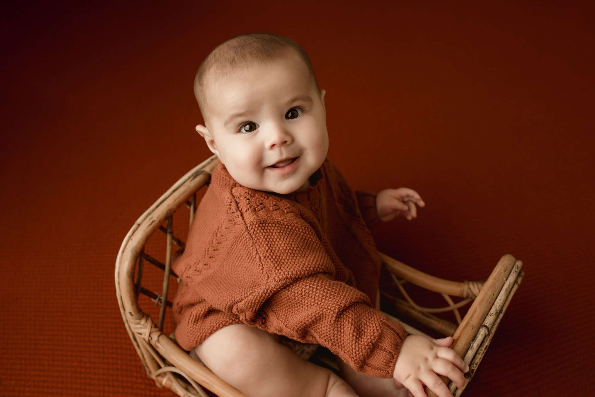 toddler sitting in a wicker basket with a orange background wearing an orange sweater smiling at the camera
